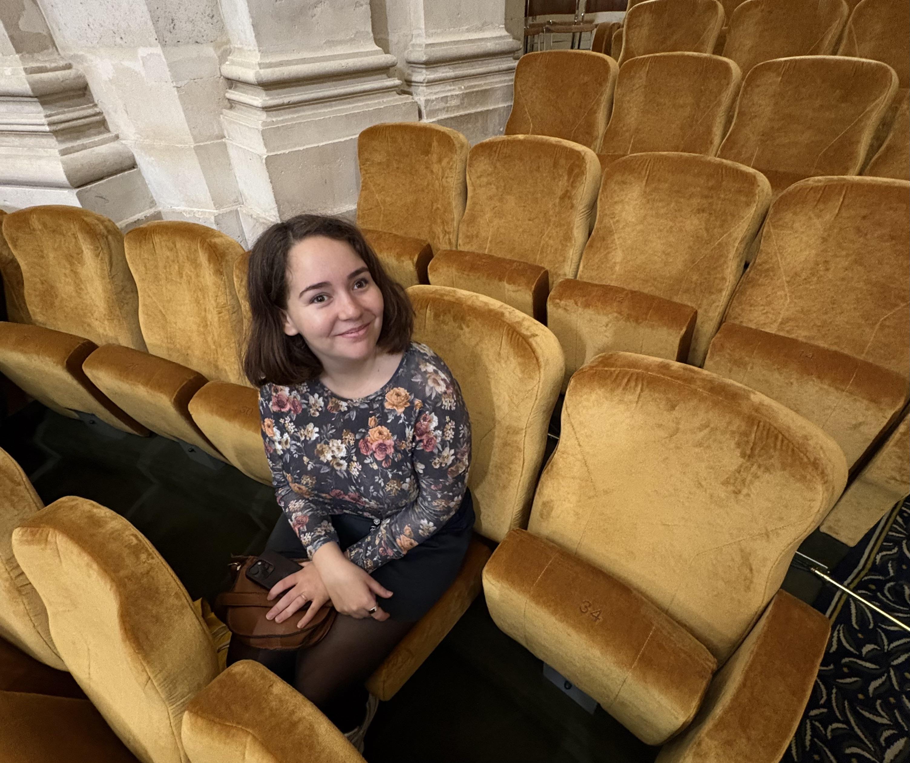 Abi, a white woman with short brown hair wearing a top with flowers and a navy blue skirt. Her hands are in her lap and she's holding a brown bag and her phone. She is sat on a golden yellow seat, surrounded more of the same seats