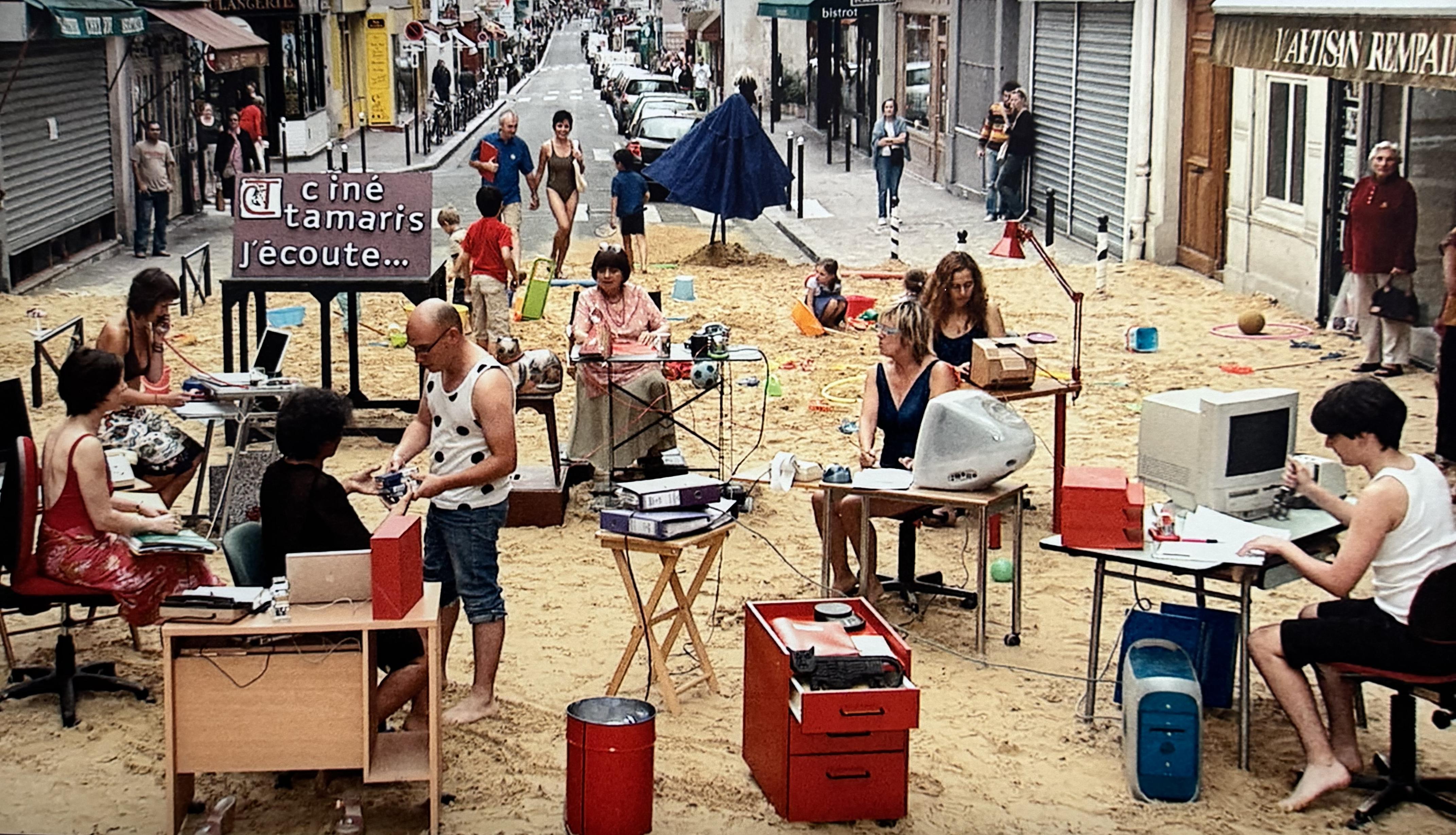 a street in Paris that has been turned into a beach. There is lots of sand on the street, with an office setup. In the background there are people doing normal beach activities