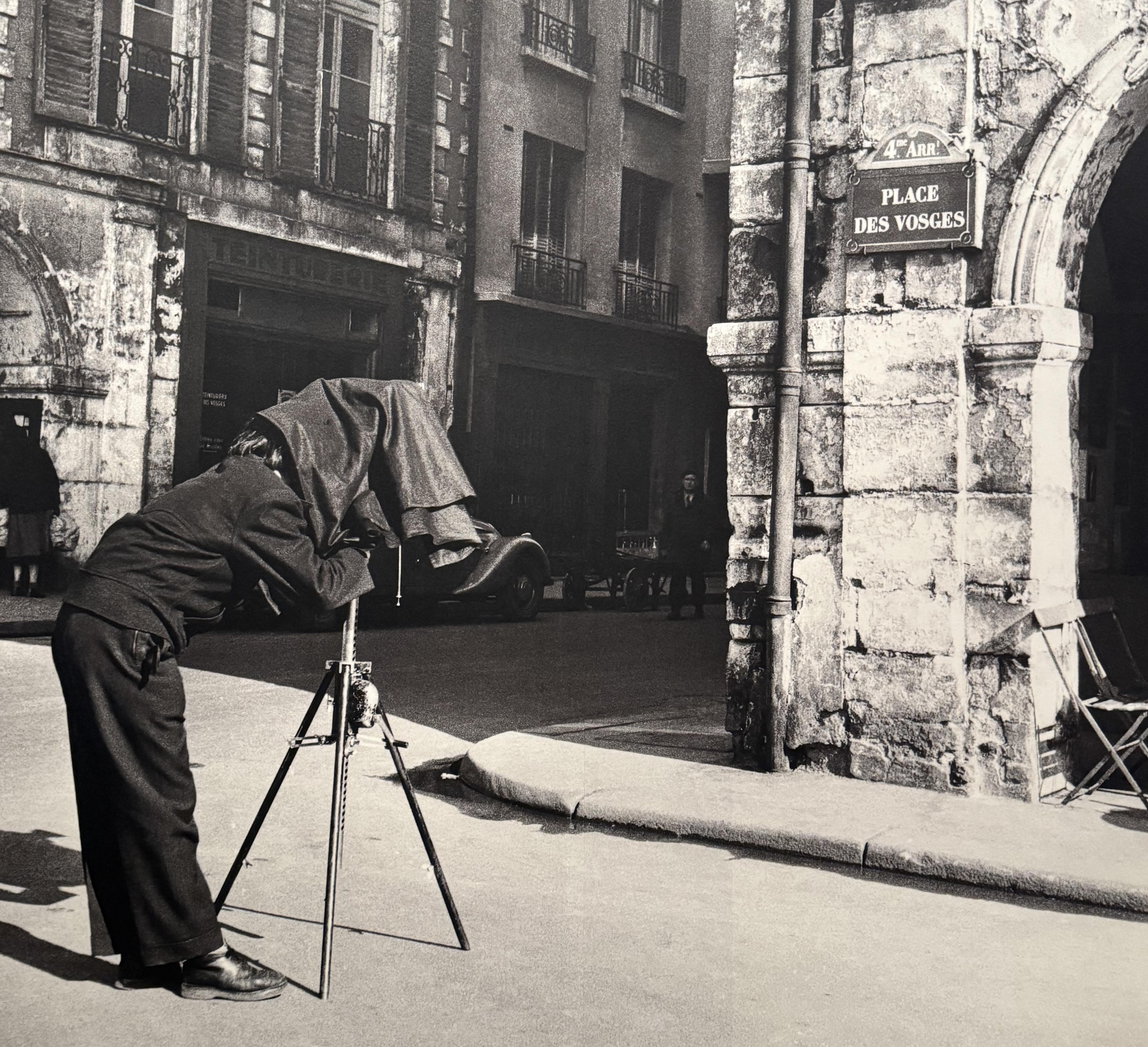 a black and white photo of Agnès Varda taking a picture at Place des Vosges. There is the street sign on the corner of a building