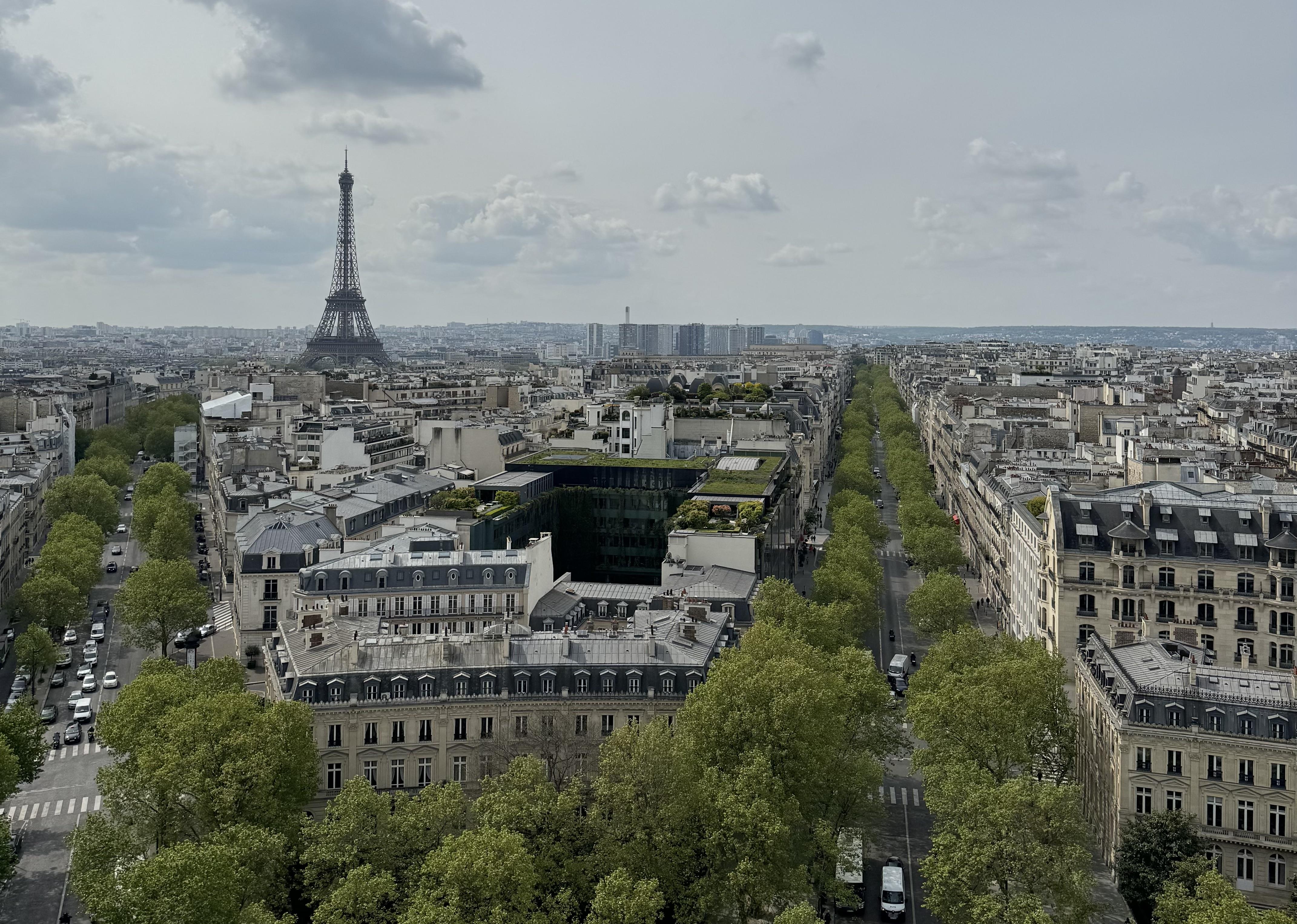 View from the top of l’Arc de Triomphe. There are two boulevards visible which are lined with trees that have green leaves. You can see the Eiffel Tower in the distance