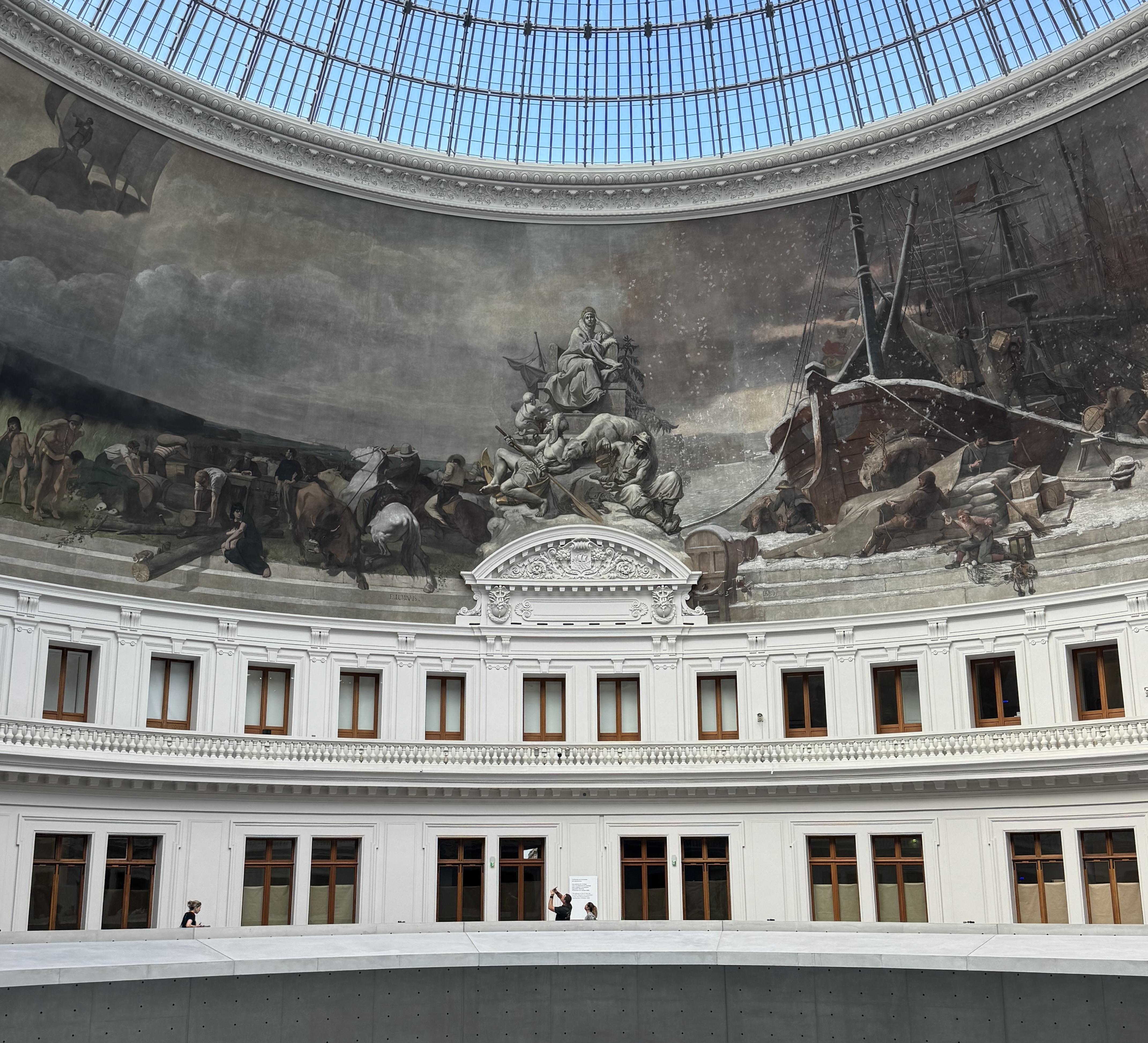 At the top, you can see the blue sky through the glass roof. In the middle there is a painting that run around the entire building. At the bottom you can see the walk way that continues and some people looking up at the painting.