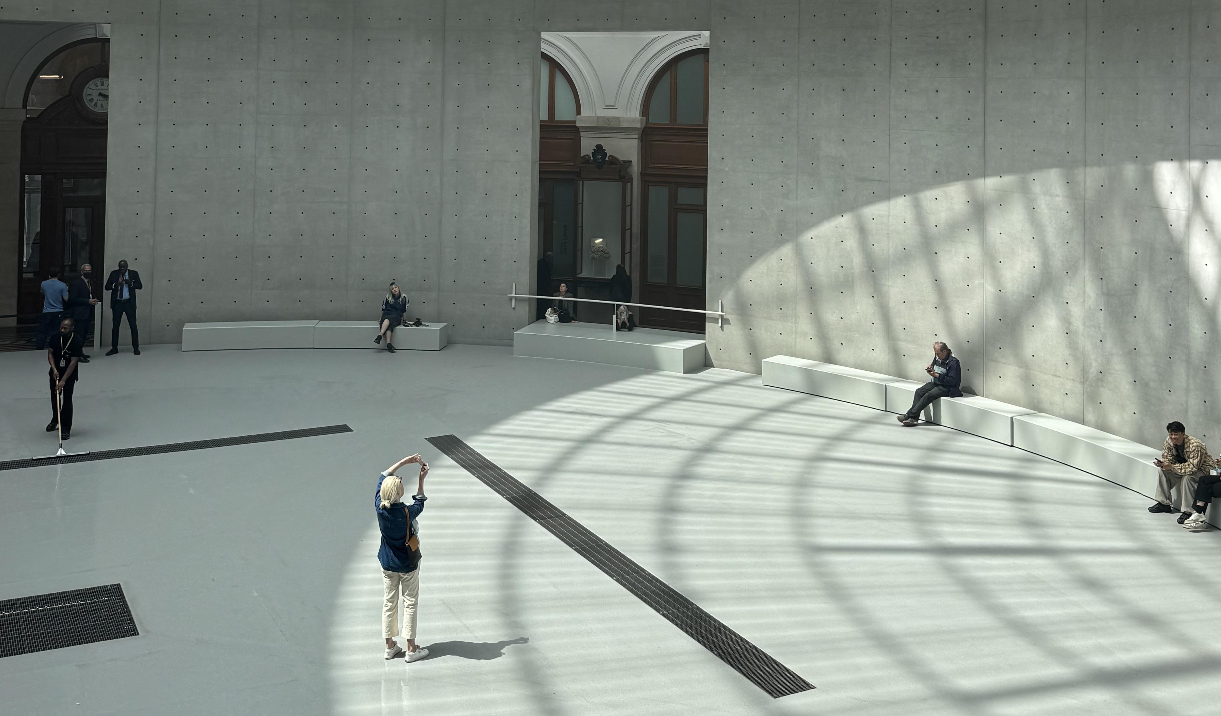 a circular room. There is no fog. There are a few people sitting around the edge of the room. There is one person standing in the centre taking a photo of the roof. The roof leaves a shadow on the floor