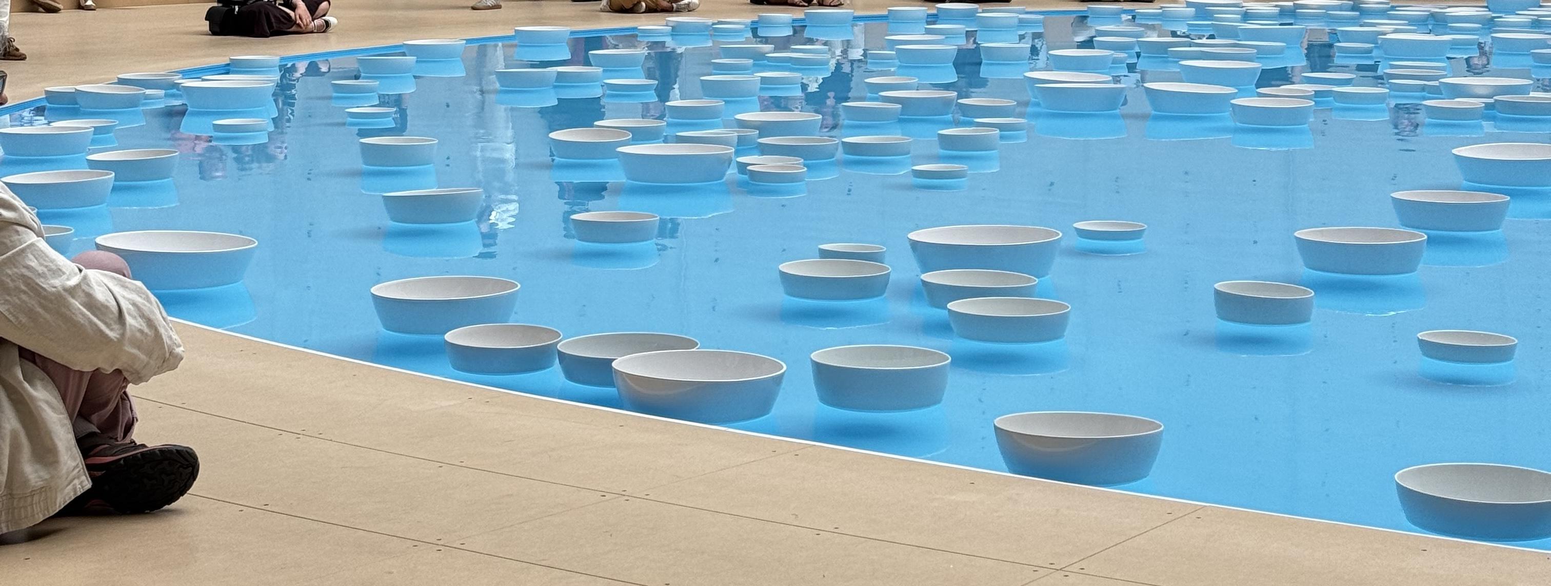 water with while bowls floating on it. The bowls vary in size. There is someone sitting close to the water on the left side. You can see some reflections of people in the water