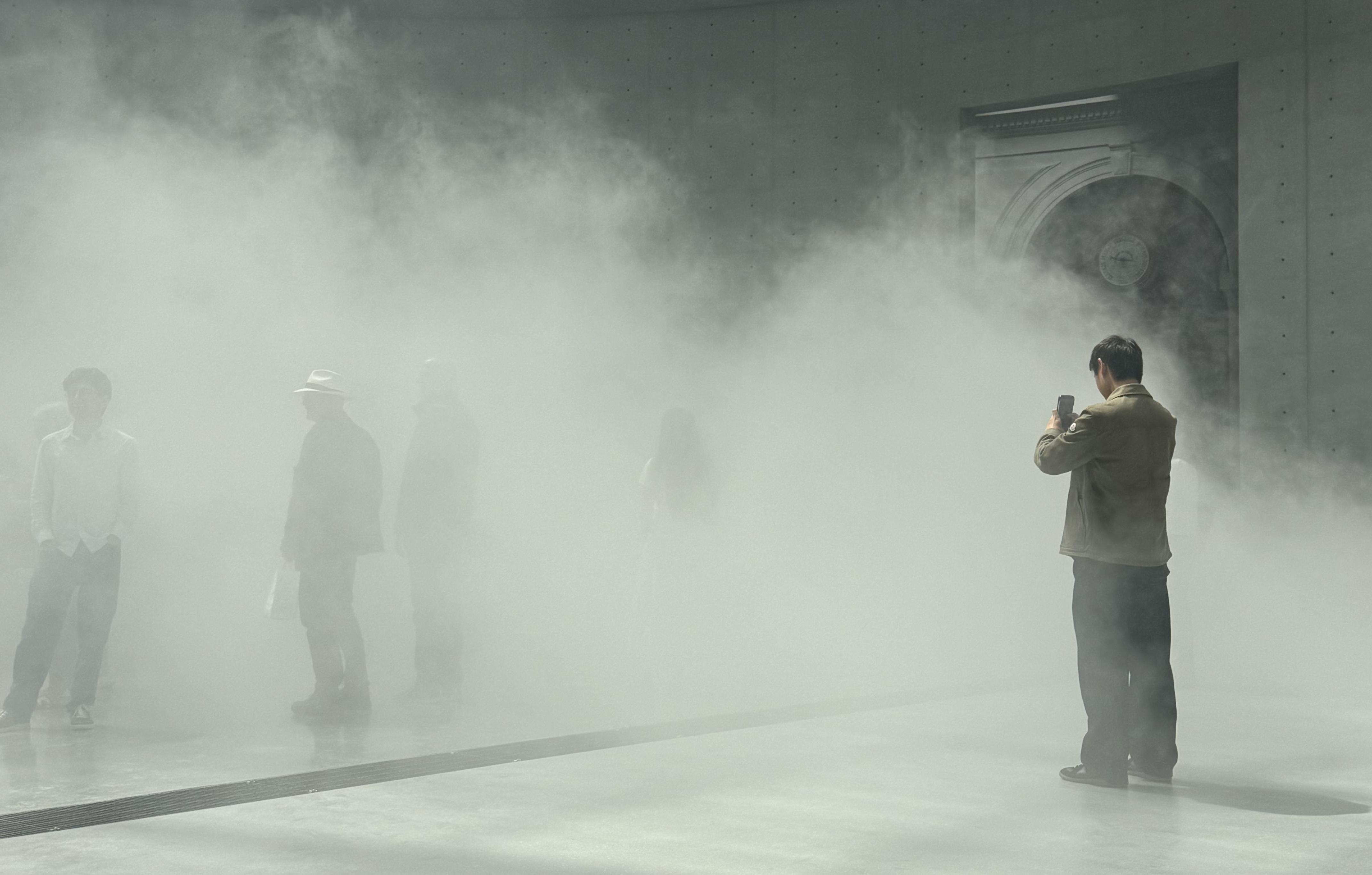 a room filled with fog. On the right, you can see one person standing in the fog. He is taking photos on his phone. In the centre of the photo you can see 5 more people who are standing in the fog so are harder to see