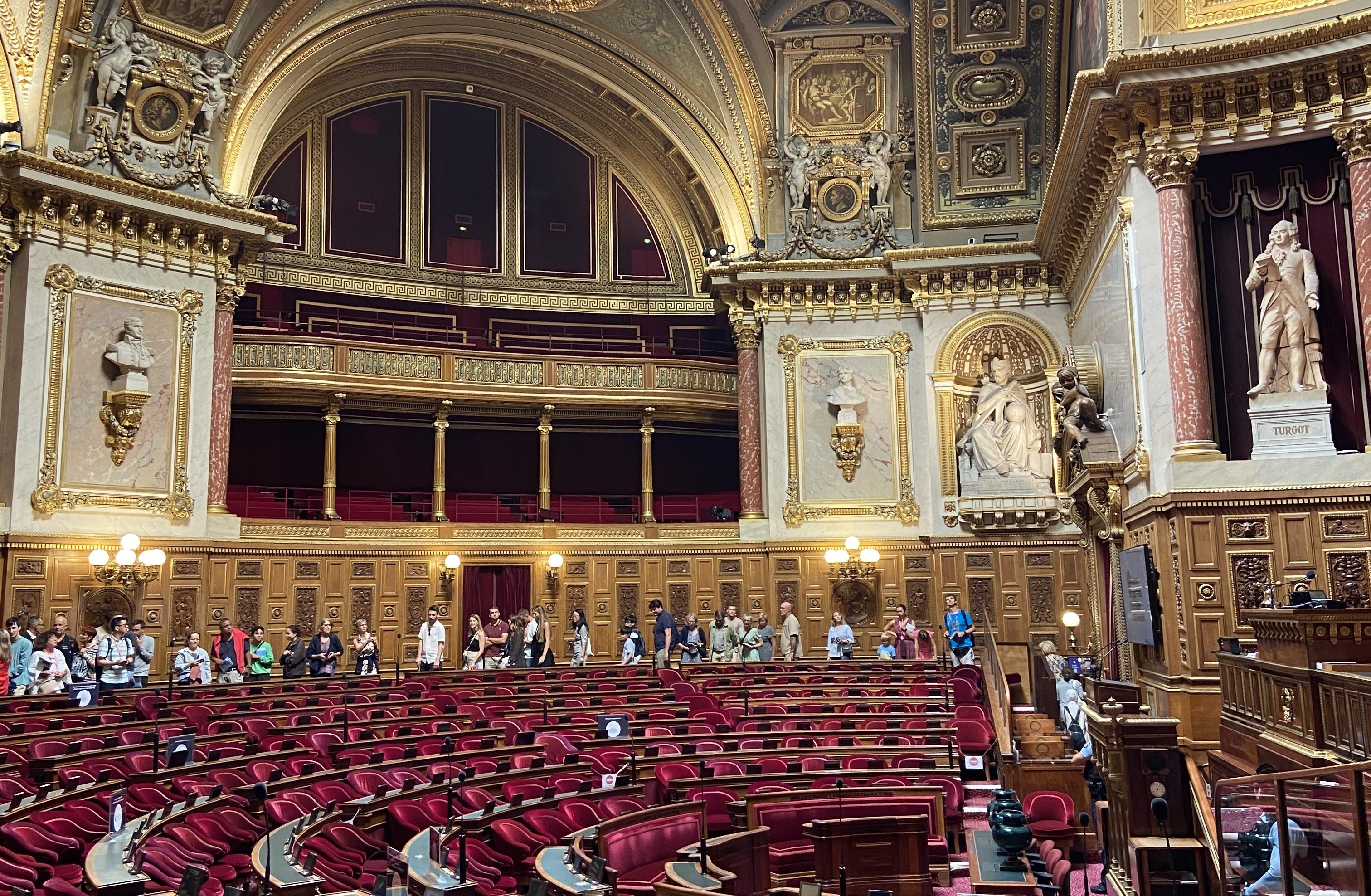 a view inside the Sénat. The walls have lots of gold details on them. The seats, positioned in a semi circle are all red. There are lots of people standing around the edge of the room