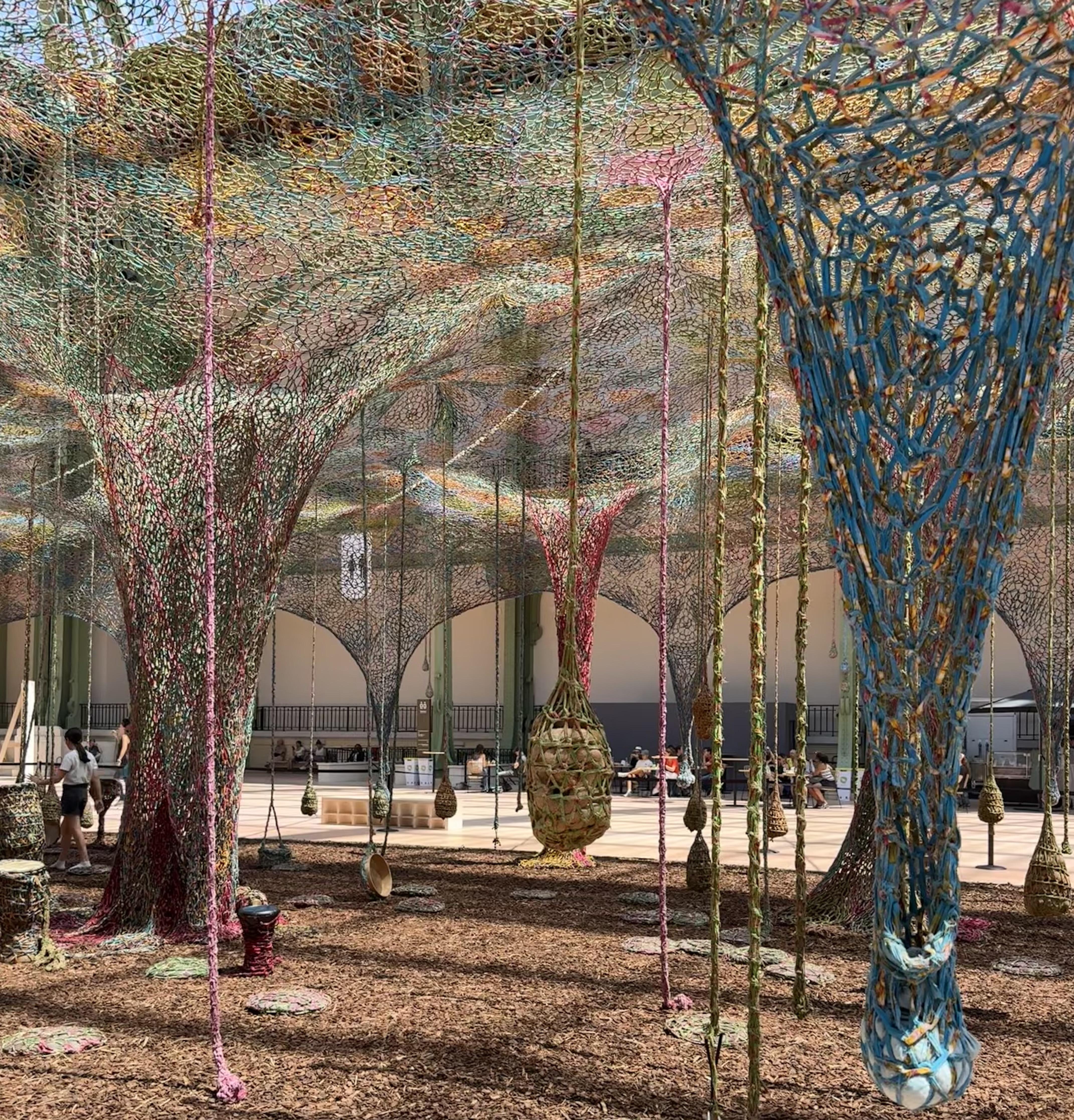 fabric that is attached to the ceiling. Each drop is of a different size and colour. There are a few drums that have been wrapped in fabric. there is bark on the ground