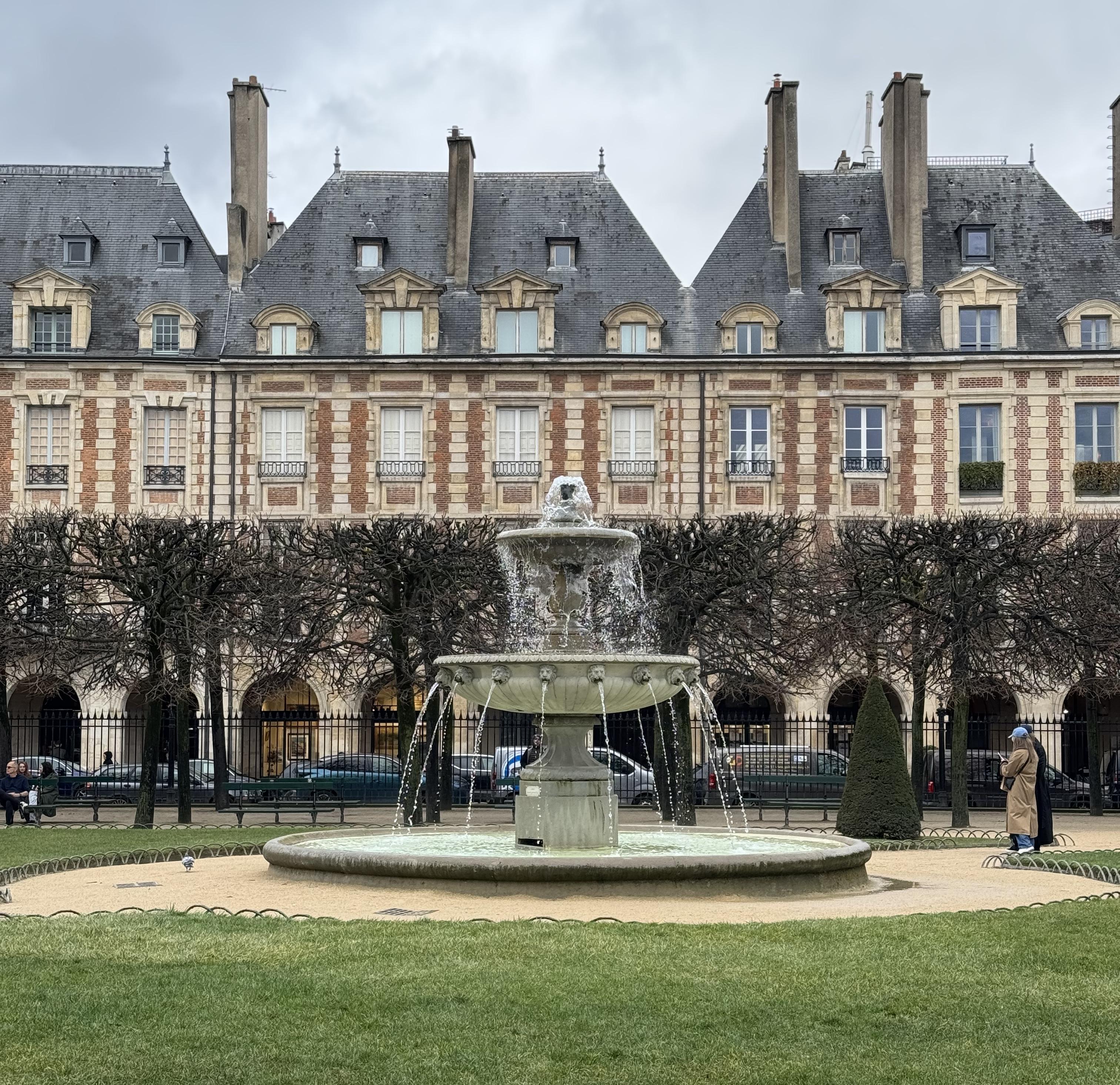 taken from inside the park, there is a fountain with water coming out of it. Behind there are some trees with no leaves. outside of the park, there is a row of buildings