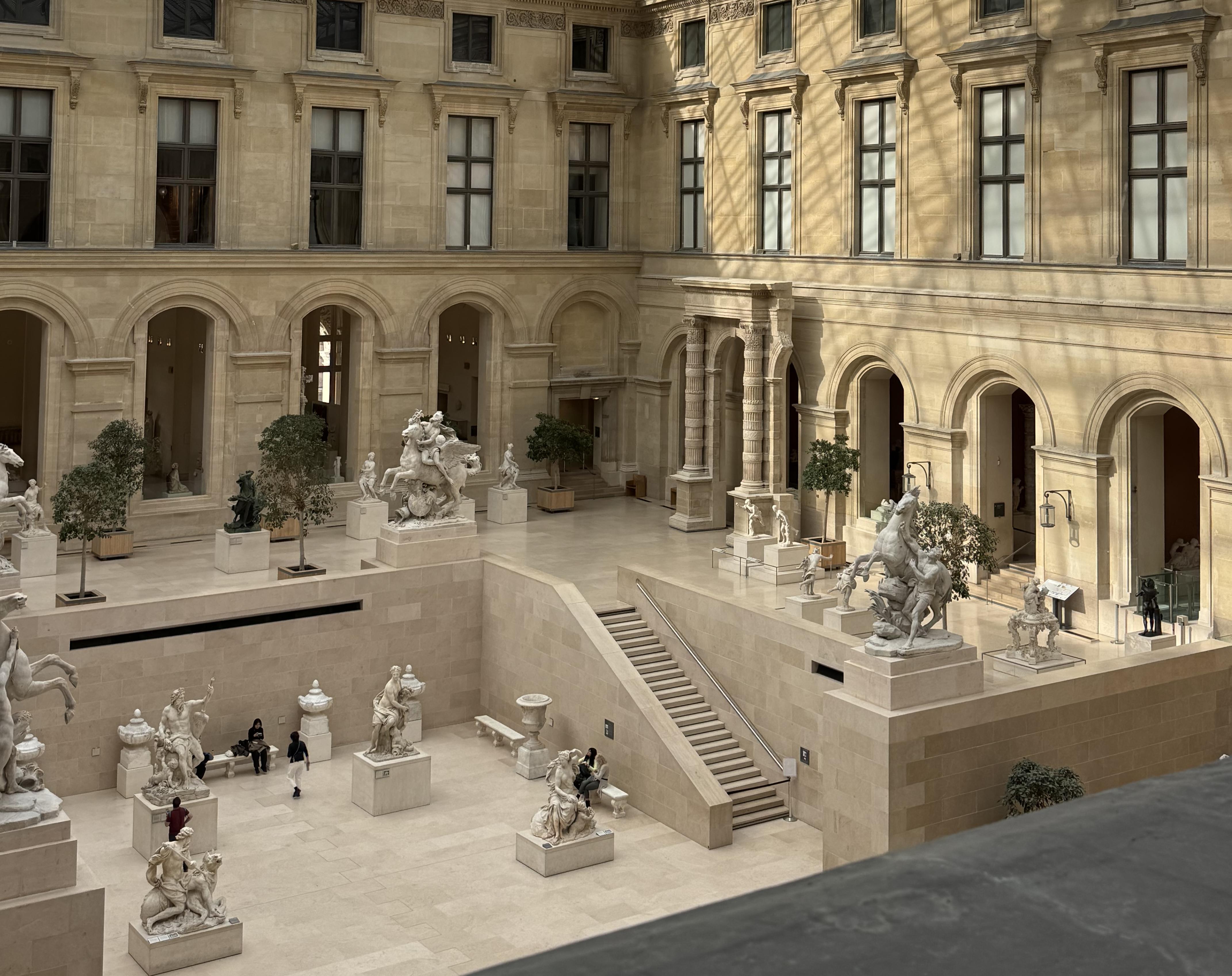 the inside of the Louvre. Over two levels, there are a handful of sculptures. On the upper level, there are some trees. On the lower level, there are five people looking at the statues. The building has a high ceiling and you can see the shadows of the roof on the walls in a grid shape.