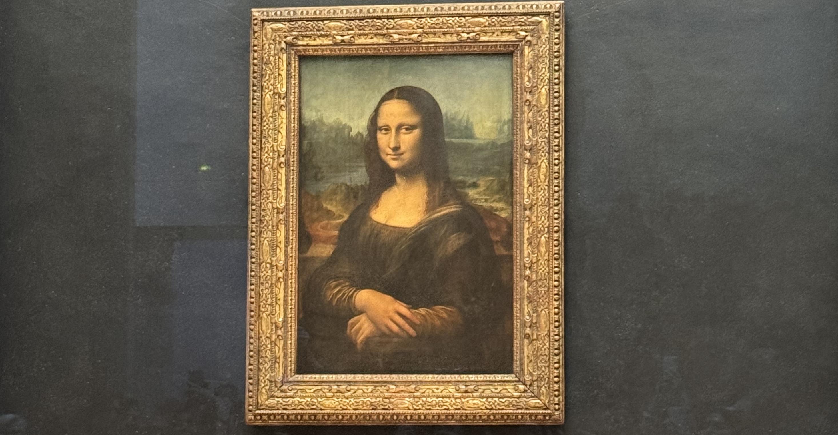 the mona lisa. the painting is behind glass, so you can see the reflection of some people. The wall that the Mona Lisa is on is a grey colour. The painting is in a gold frame