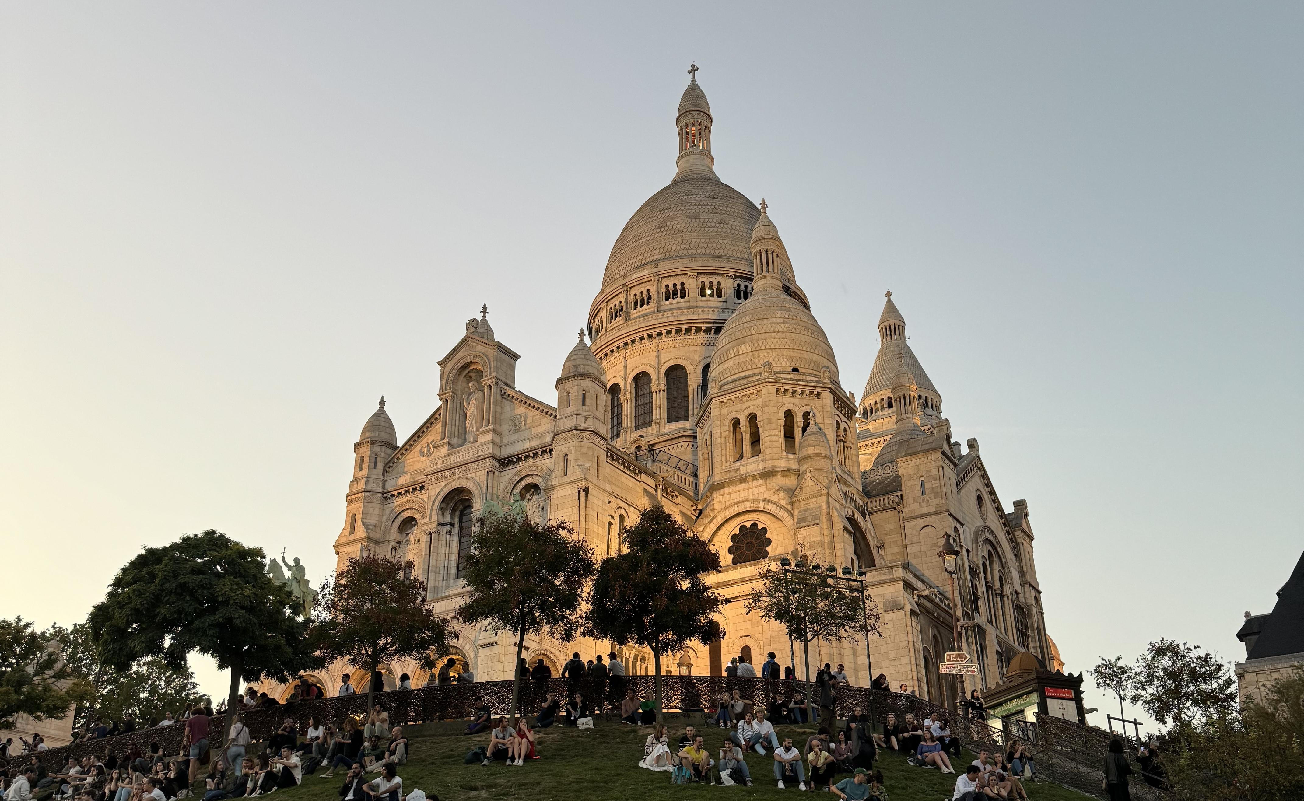 the sacre cœur at sunset, it has a orange glow. There are people sitting on the grass bank infront of it