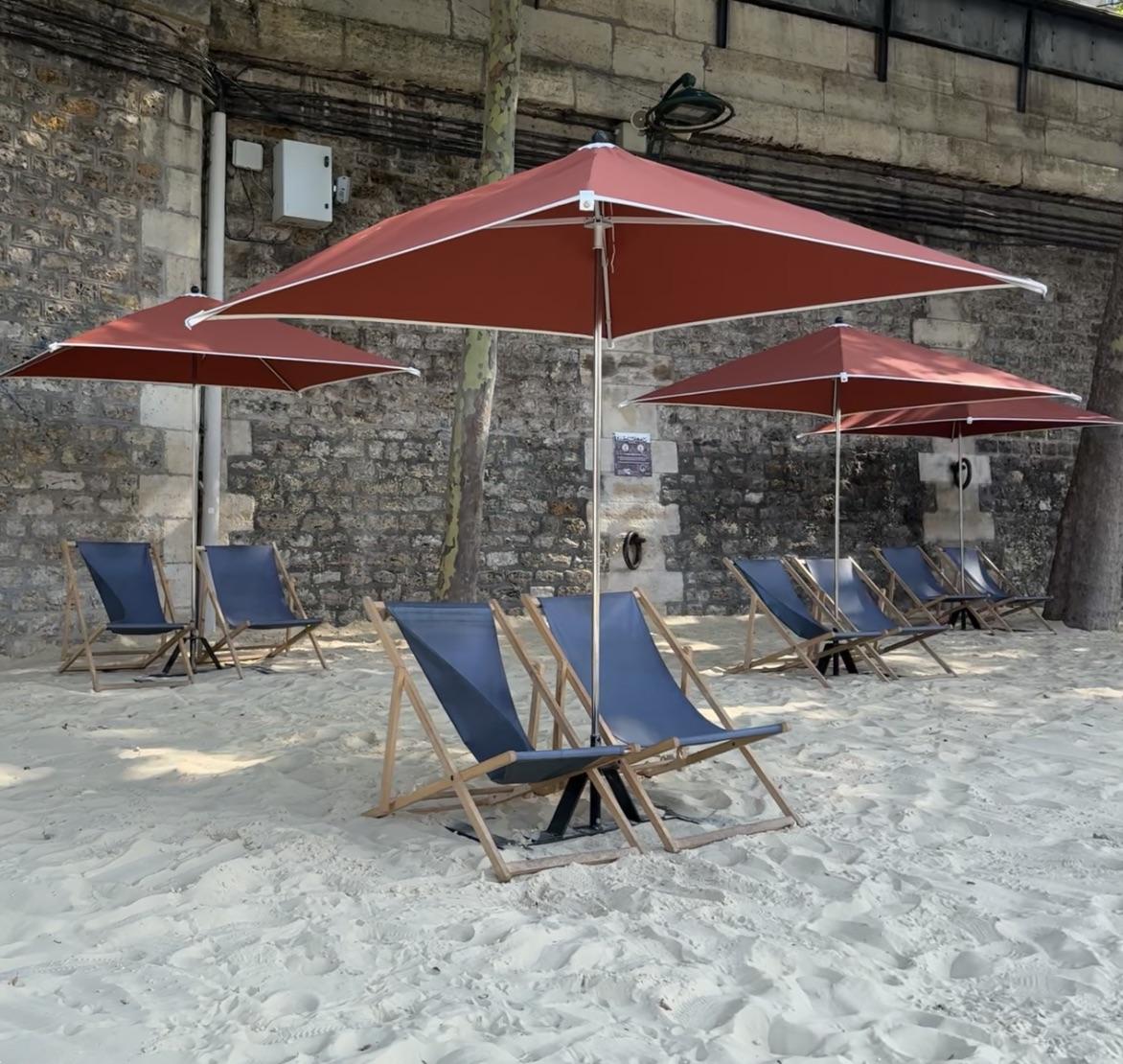 four sets of two blue deckchairs. The are on sand. Each set of deckchairs has a red parasol with them