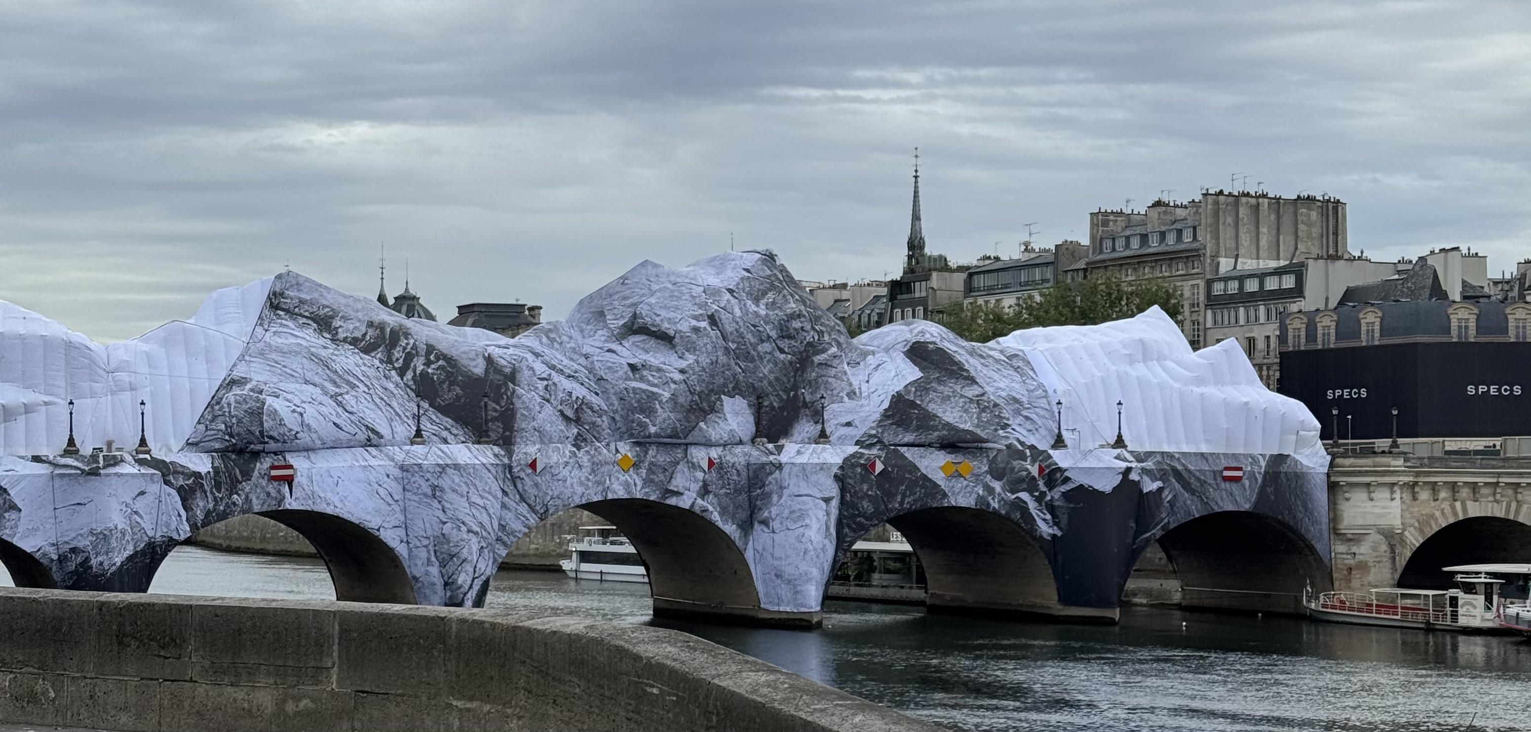 Pont Neuf wrapped with a fabric that looks like mountains. The left side and the right side have been damaged by the weather. You can see the bright white under layer. Behind the bridge on the right, there is an advertisement which says SPECS.