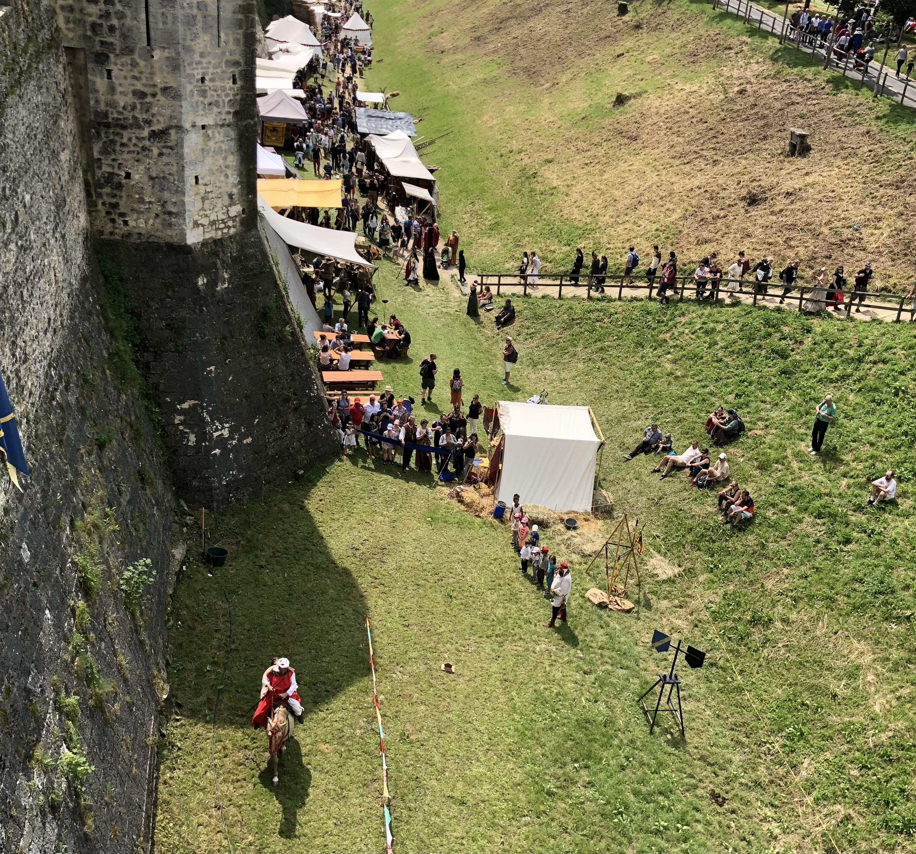 lots of people standing outside the city walls while visiting the medieval festival. There is someone riding a horse