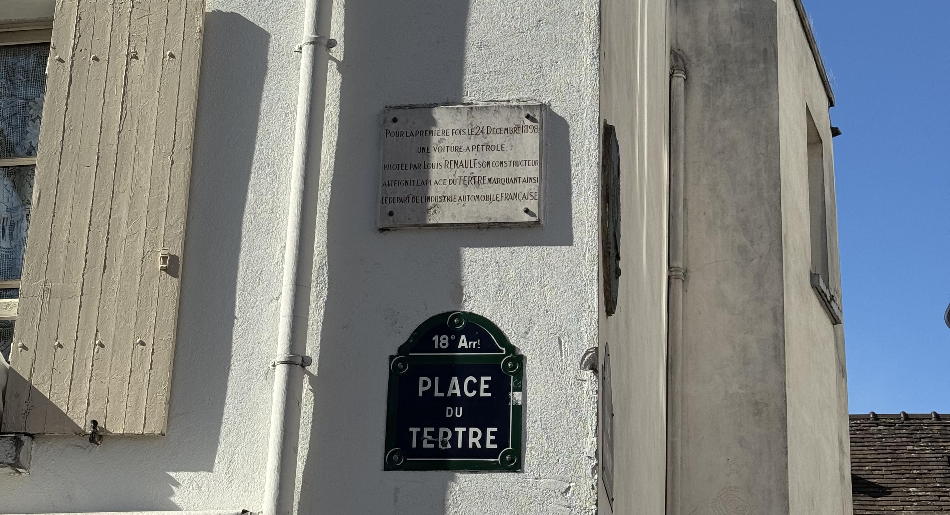 a white plaques on the wall reading "pour la premiere fois le 24 decembre 1898, une voiture a petrole pilotee par louis renault son constructeur atteignit la place du tertre marquant ainsi le depart de l'industrie automobile francaise". Underneath you can see the street sign in the typical blue Paris style: 18arr, place du tertre