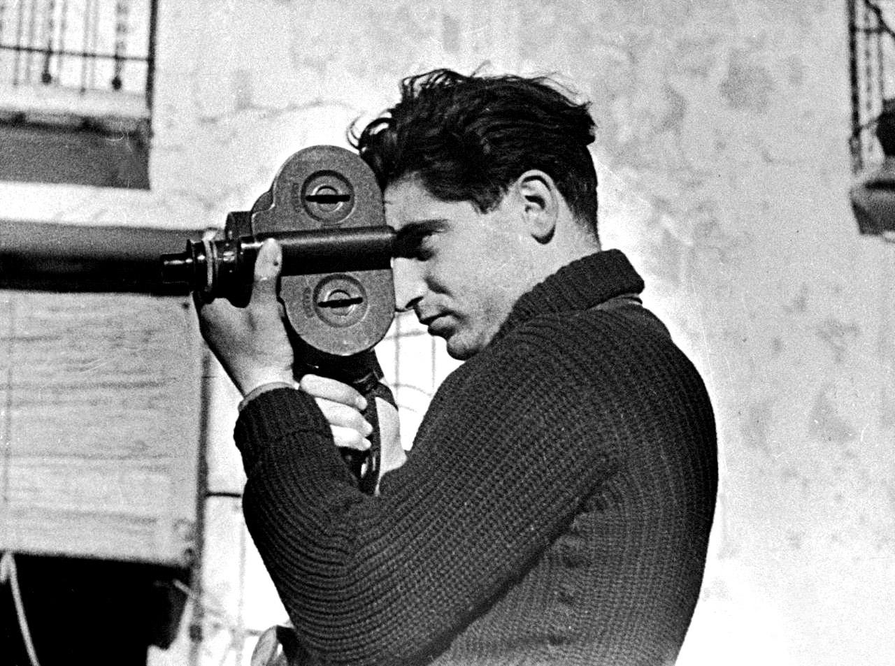 a black and white photo of Robert Capa. He is looking to the left while holding a camera