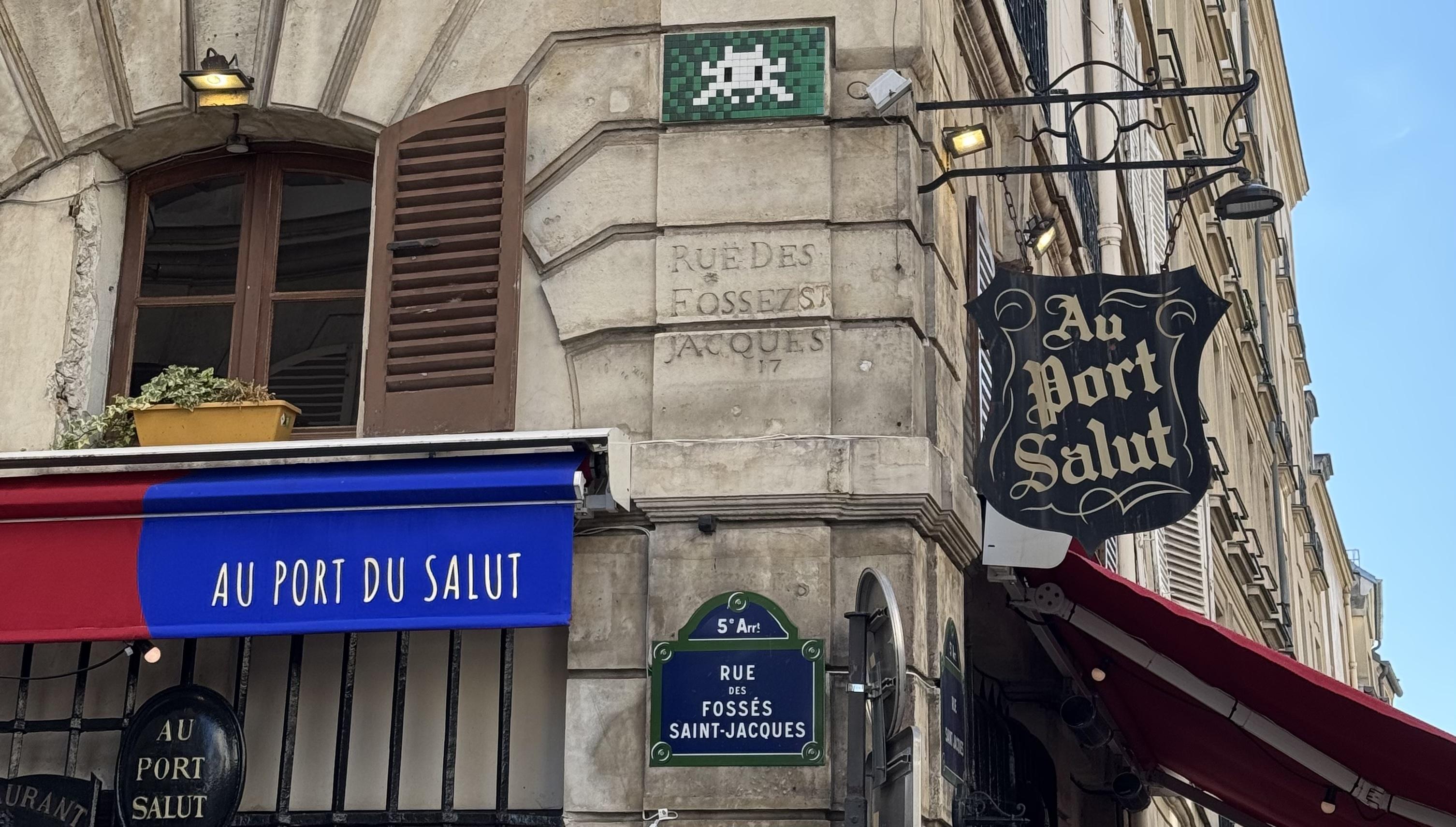 a street sign in Paris for the street 'rue des fosses saint jacques'. In the middle there is the only street sign that is carved into stone, and at the bottom there is the modern day sign - a blue rectangle with a green border with white text. At the top, attached to the rectangle there is a semi circle which says which arrondissement you are in. You can also see a space invader at the top, and the the restaurant name Au Port du Salut