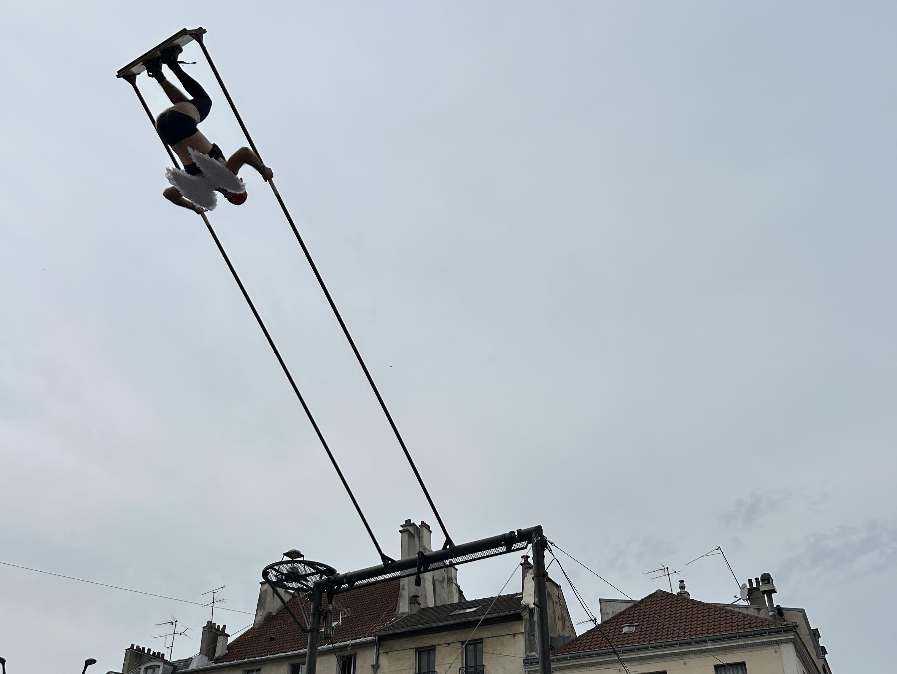 a man upside down on a standing swing. He is wearing while angel wings. You can see the top of the houses