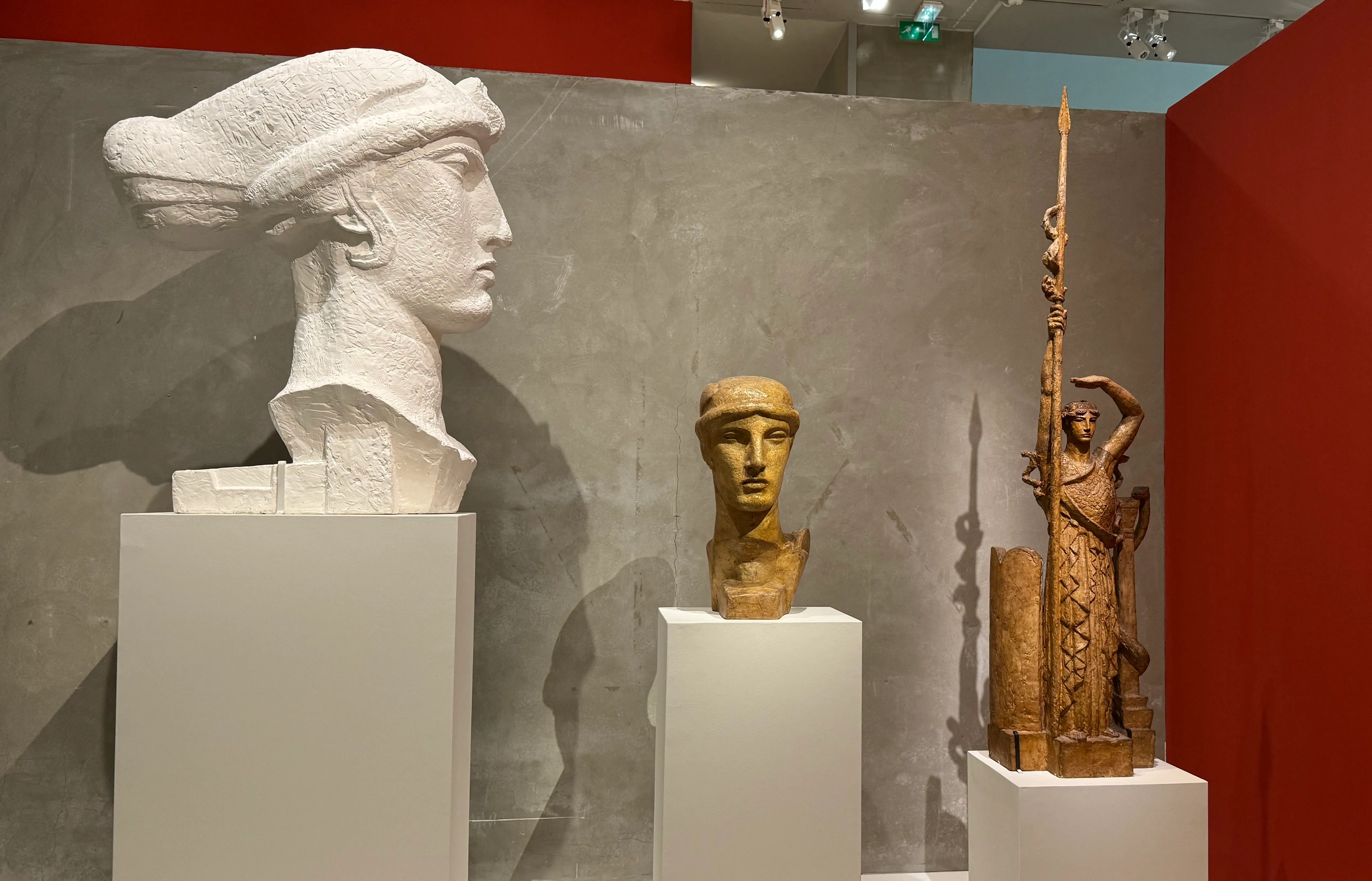 three sculptures. The sculpture on the left is a woman looking right made of plaster. The middle sculpture is lower, and is facing towards us. It&#x27;s made of plaster but has a coating that looks gold. On the right, we have the third sculpture which is facing towards us, this sculpture is a woman standing tall, with one hand holding a long spear, and the other hand above her head