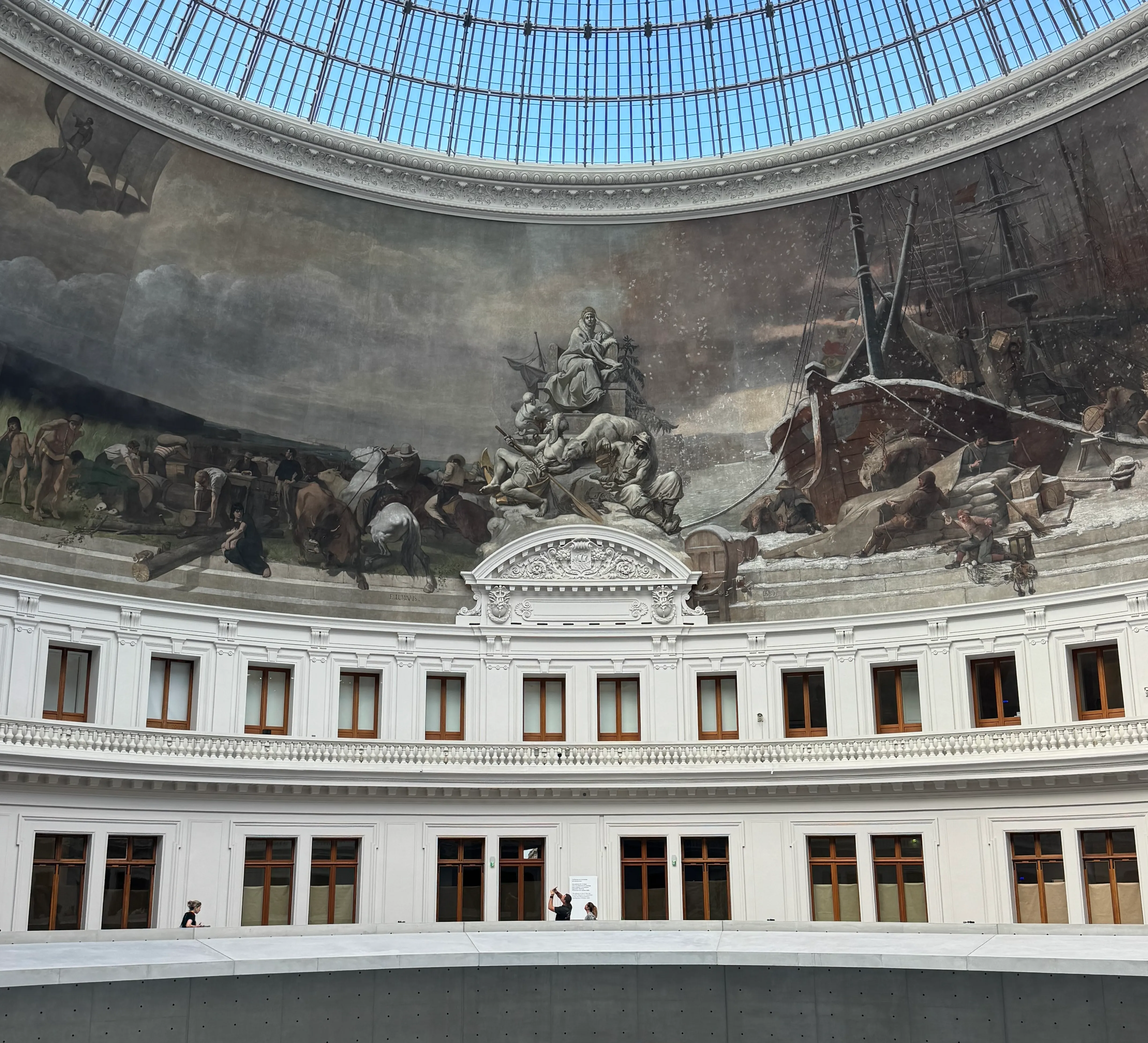 At the top, you can see the blue sky through the glass roof. In the middle there is a painting that run around the entire building. At the bottom you can see the walk way that continues and some people looking up at the painting.