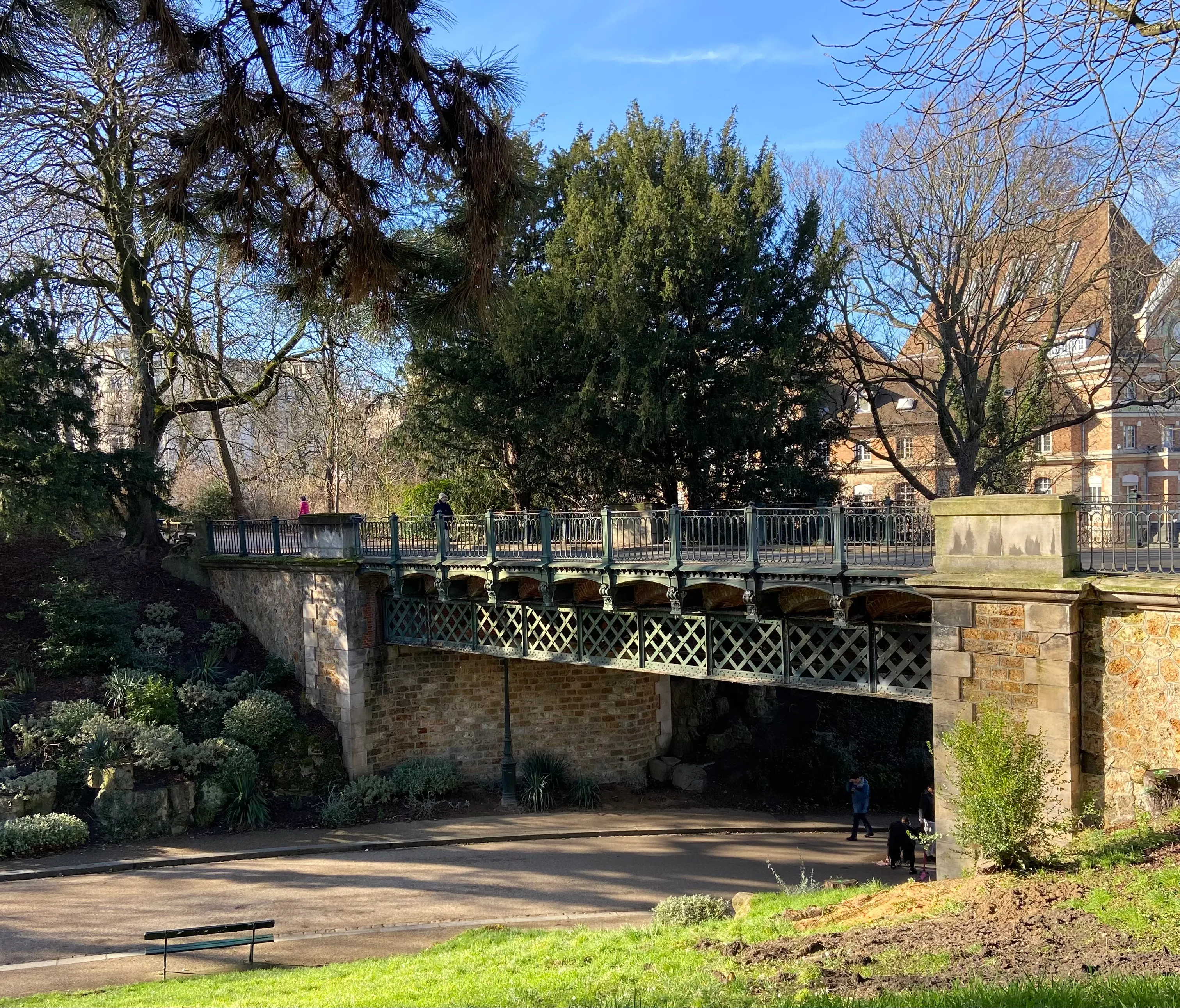 parc des Buttes-Chaumont. A bridge goes over a pedestrian walkway. You can see some brick buildings in the back.