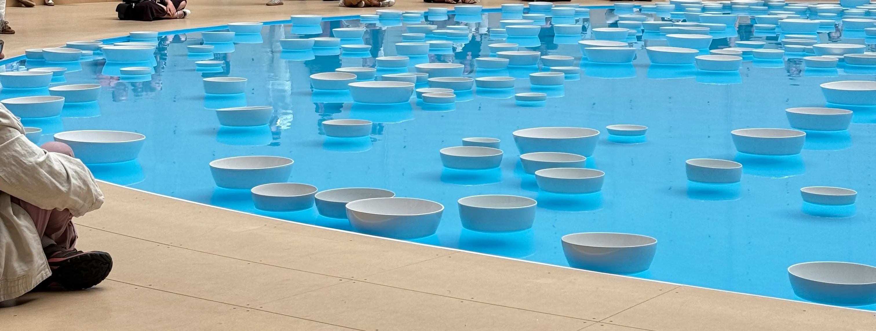 water with while bowls floating on it. The bowls vary in size. There is someone sitting close to the water on the left side. You can see some reflections of people in the water