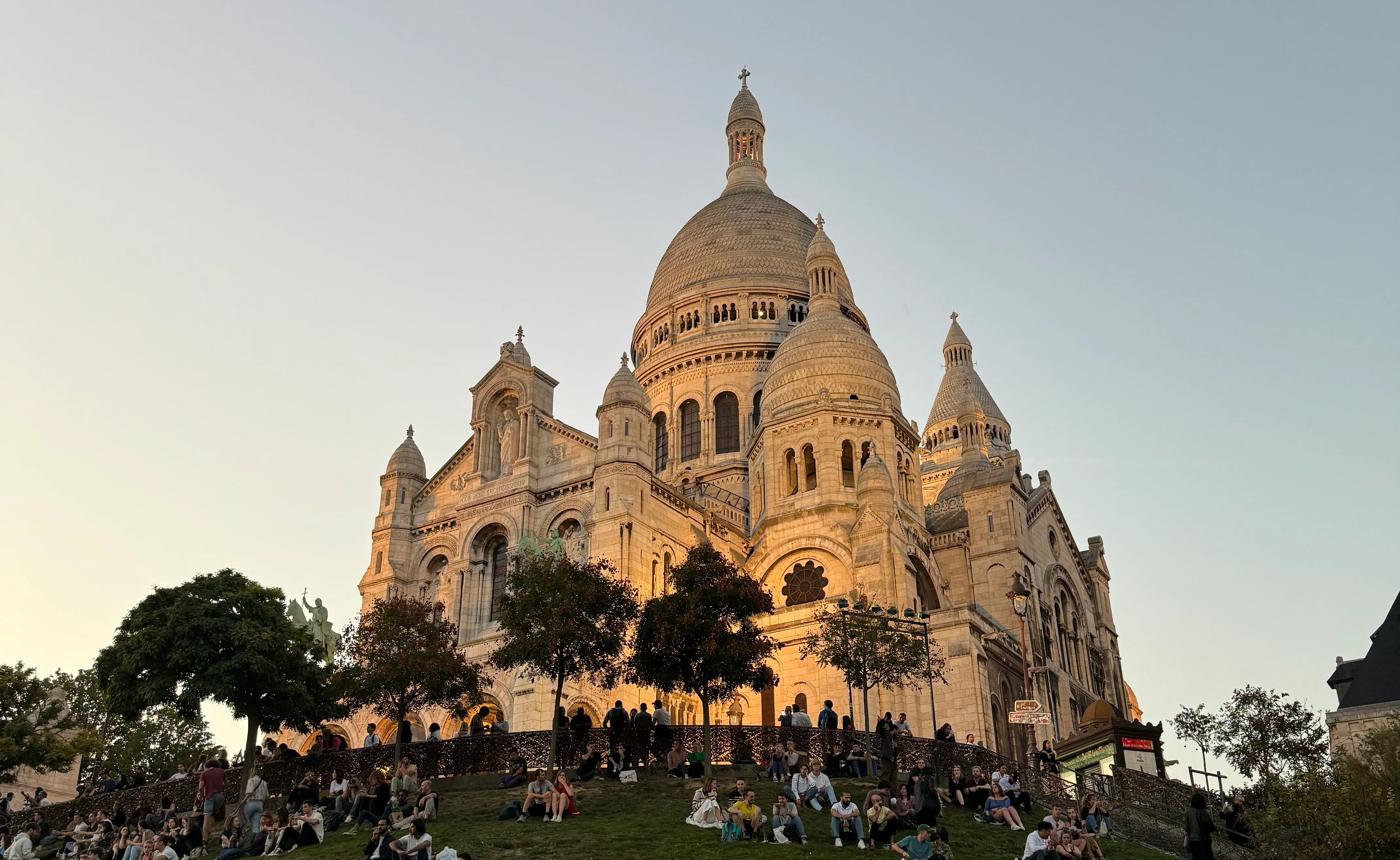 the sacre cœur at sunset, it has a orange glow. There are people sitting on the grass bank infront of it