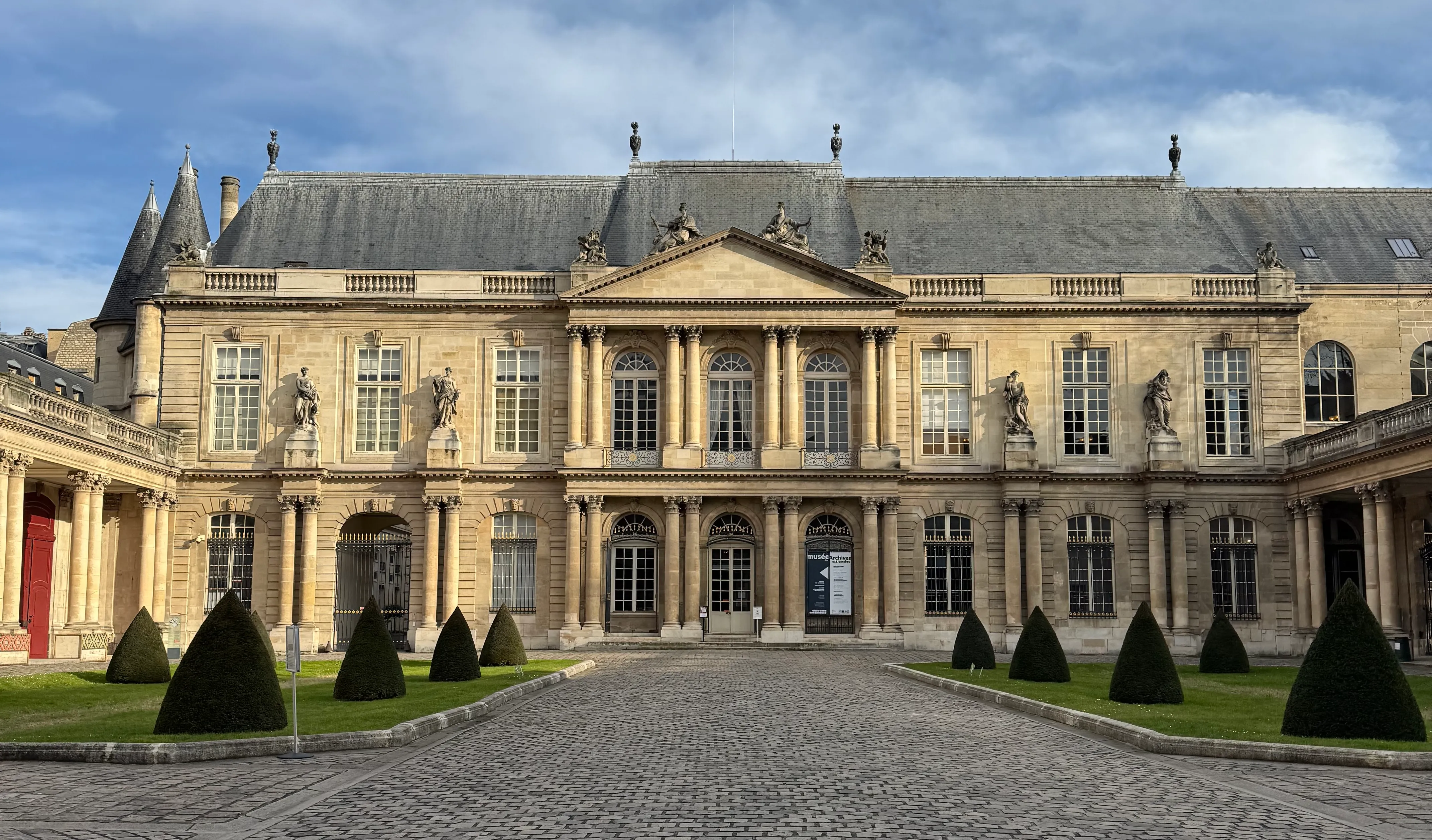 the building that houses the national archives. In front of the doors there are two grass patches that have shrubs in the shape of a cone