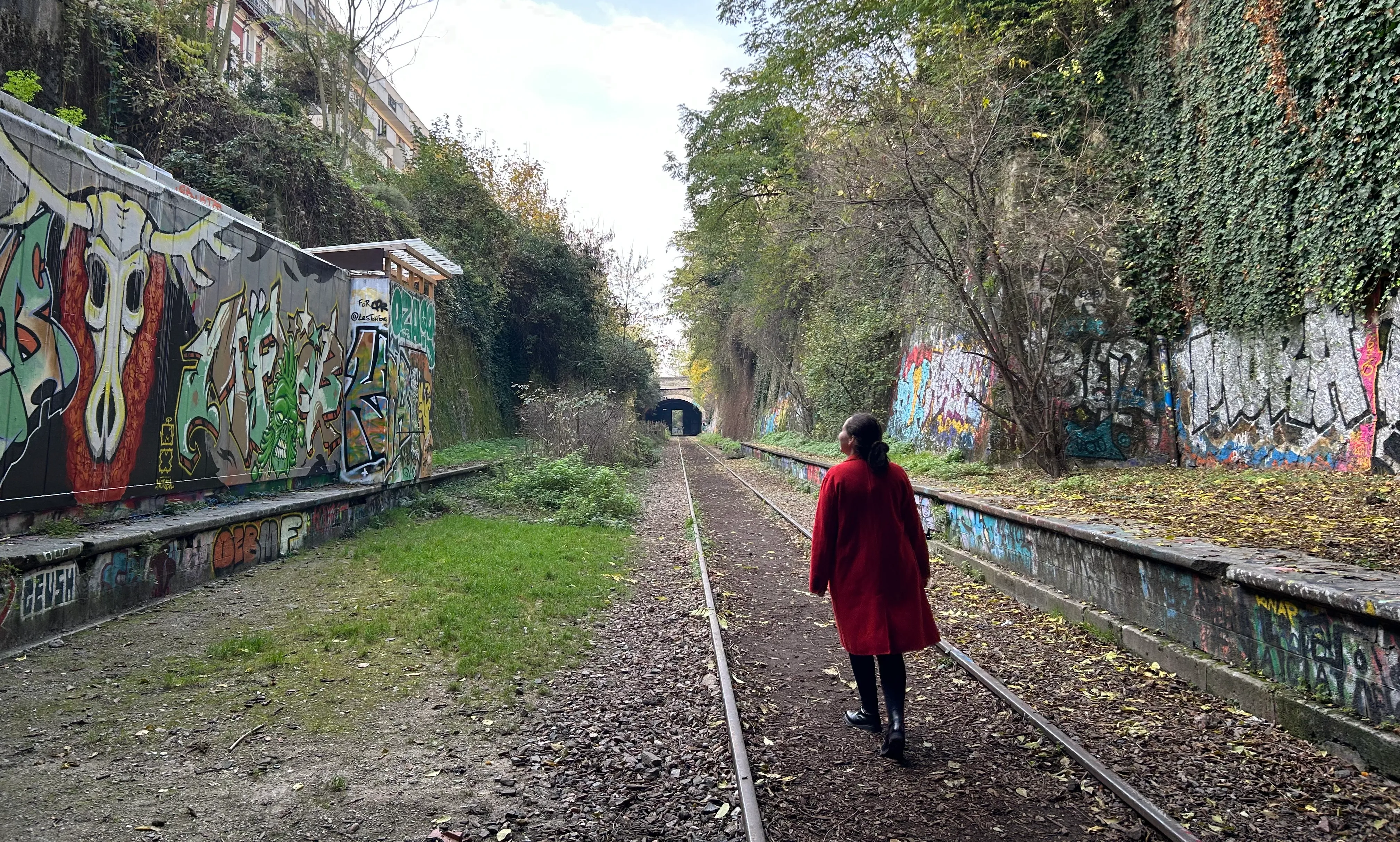 a woman wearing a red jacket, walking along the old train track with the station platform on either side. there is a bridge in the distance that the track goes under