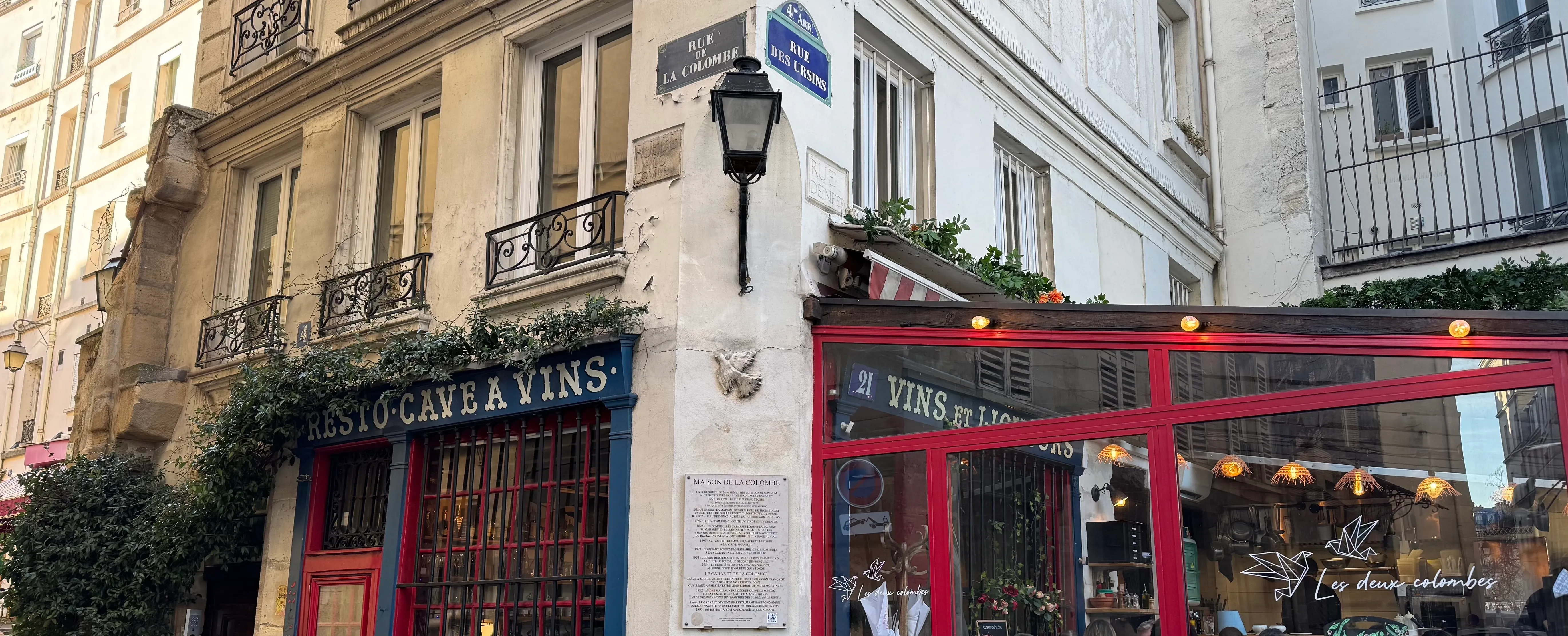 A restaurant called &#x27;les deux colombes&#x27;. on the wall there is a plaque that explains the history, above this there is a stone dove. You can see the street names carved into the stone along with the modern day signs.